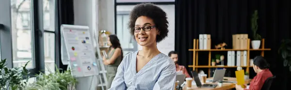 Gioiosa donna afro-americana impacchettare i suoi oggetti durante licenziamento, colleghi sullo sfondo. — Foto stock