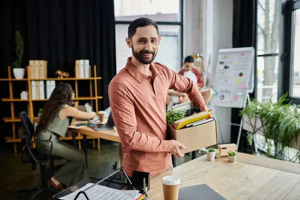 Stylish man packing his belongings during lay off in modern office, colleagues on backdrop. — Stock Photo