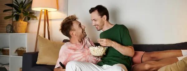Two men share popcorn and laughter while snuggling on the couch together. — Stock Photo
