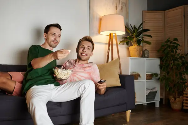 Two men share a joyful moment on their couch, enjoying popcorn and each other. — Stock Photo