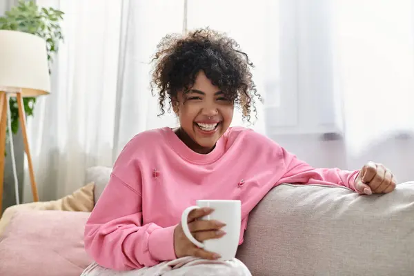 A young woman smiles brightly while holding a mug in her cozy living room. — Stock Photo