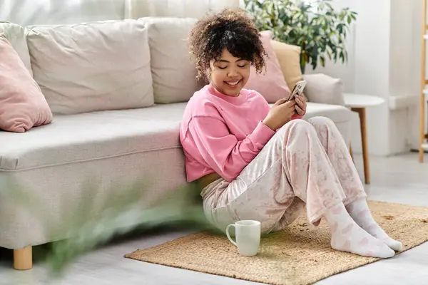A young woman in pink loungewear smiles while using her phone at home. — Stock Photo
