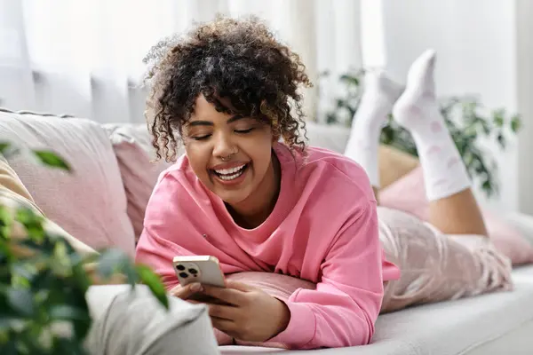 A cheerful woman relaxes on the couch, happily engaging with her mobile device. — Stock Photo