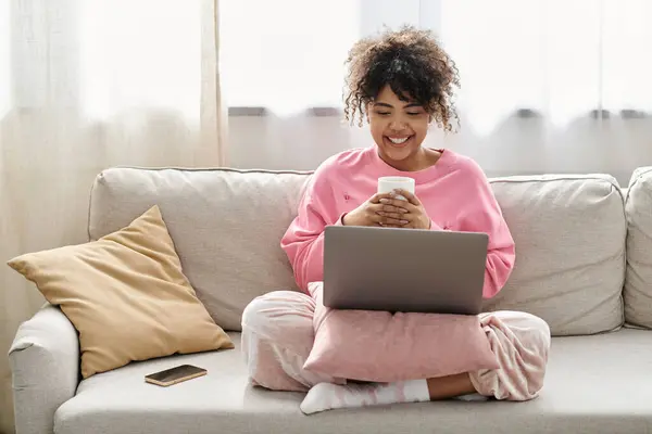 A young woman smiles while using her laptop and sipping coffee in a cozy setting. — Stock Photo