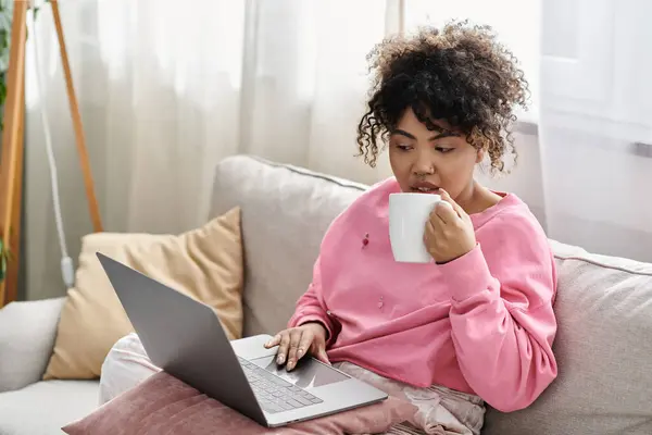 A young woman sips coffee while focused on her laptop at home. — Stock Photo