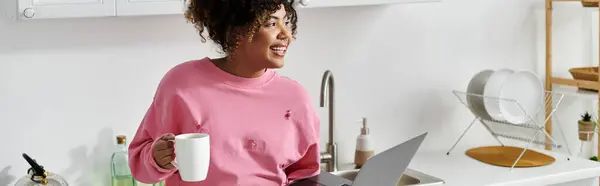 Smiling woman sipping coffee while working on her laptop in a bright kitchen. — Stock Photo