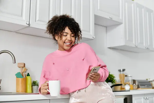 Mulher sorridente relaxa com café enquanto usa seu telefone em uma cozinha brilhante. — Fotografia de Stock