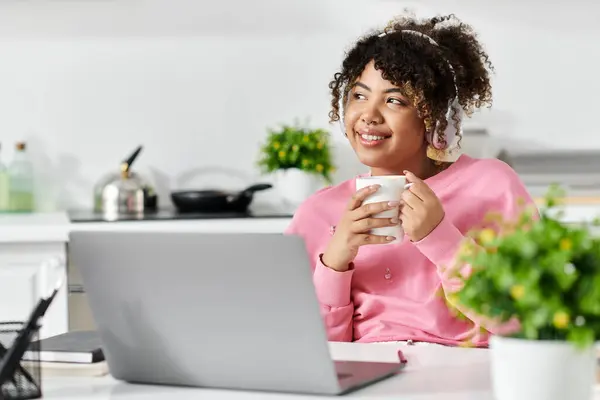 Una mujer joven se relaja en casa, bebiendo una bebida caliente mientras trabaja en su computadora portátil. - foto de stock