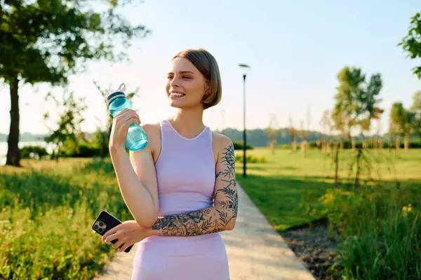 A joyful young woman relaxes in a sunny park, sipping water. — Stock Photo