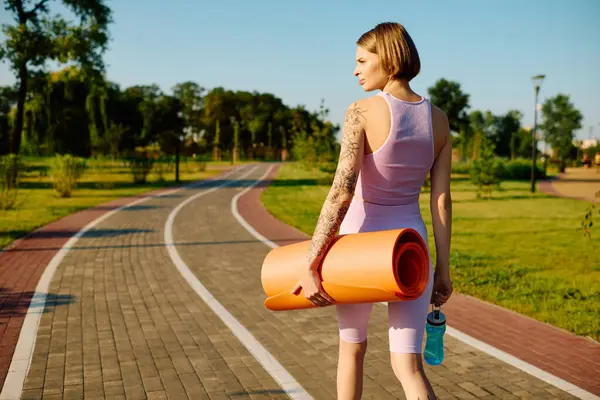 Uma mulher carrega tapetes de ioga e água em um ambiente de parque sereno. — Fotografia de Stock