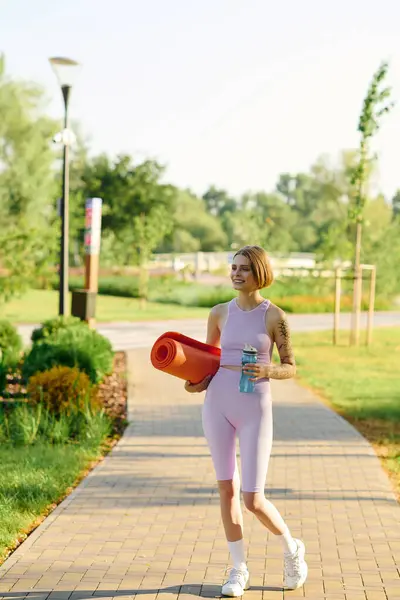 Cheerful woman strolls through a vibrant park with a yoga mat and refreshing drink. — Stock Photo