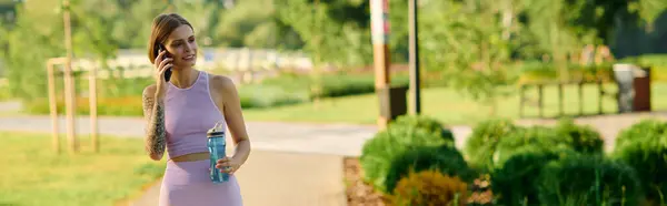 A woman in activewear strolls through a park, sipping water and on a phone call. — Stock Photo