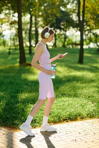 She walks through a vibrant park, headphones in, sipping water and engaged on her phone. — Stock Photo