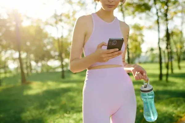Una giovane donna sta alla luce del sole, si concentra sul telefono e tiene in mano una bottiglia d'acqua. — Foto stock