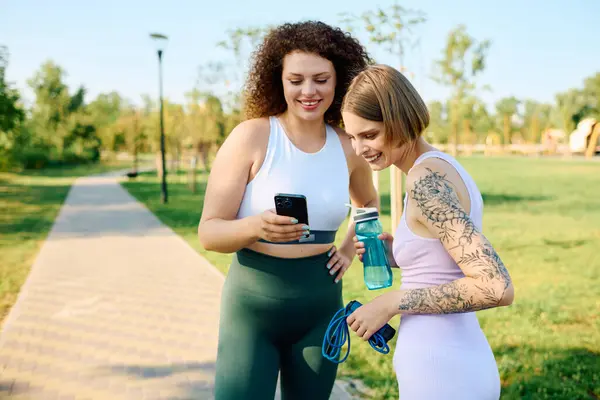 Zwei Frauen lachen zusammen, während sie in einem sonnigen Park ihre Telefone checken. — Stockfoto