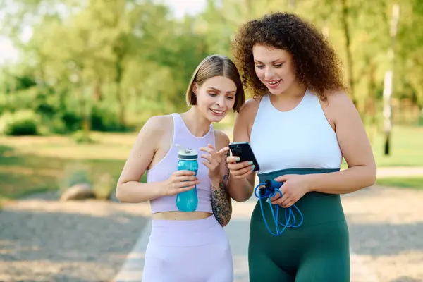 Two women share a joyful moment together in the park, engaging with their phones. — Stock Photo