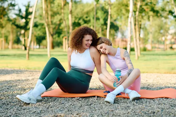 Duas mulheres compartilham risos e carinho enquanto se sentam em tapetes de ioga no parque. — Fotografia de Stock