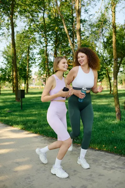 Duas mulheres que jogging feliz lado a lado em um ajuste vibrante do parque. — Fotografia de Stock