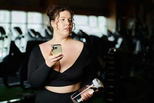 Uma mulher plus-size segura seu telefone e garrafa de água, concentrando-se em sua jornada de fitness no ginásio. — Fotografia de Stock