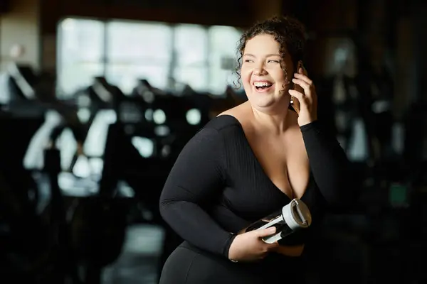 Una giovane donna irradia gioia mentre chatta al telefono, abbracciando la positività del corpo in palestra. — Foto stock