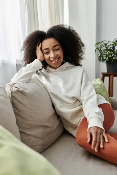 Una giovane donna dai capelli ricci si rilassa comodamente a casa, sorridendo nel suo accogliente abbigliamento sul divano. — Foto stock