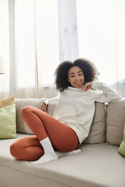 A young woman enjoys a cozy moment at home, lounging comfortably on her couch in a warm outfit. — Stock Photo