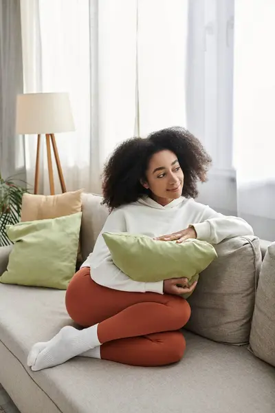 A young woman relaxes on a sofa, holding a pillow and wrapped in her cozy outfit during a serene moment at home. — Stock Photo