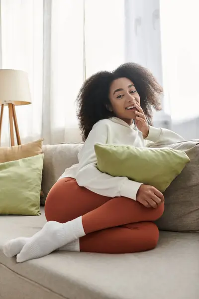 A young woman relaxes on the couch, smiling while surrounded by soft pillows in her cozy living room. — Stock Photo
