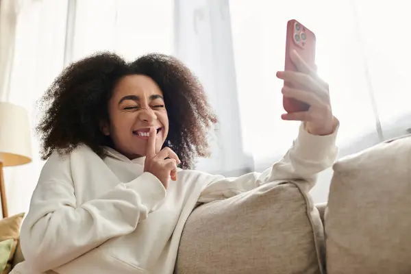 A young woman with curly hair smiles playfully at her phone while lounging comfortably at home. — Stock Photo
