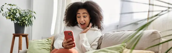 Une jeune femme aux cheveux bouclés se détend sur le canapé, surprise par quelque chose sur son téléphone. — Photo de stock