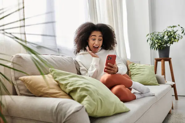 Une jeune femme dans une tenue confortable sourit tout en s'engageant avec son téléphone dans son salon. — Photo de stock