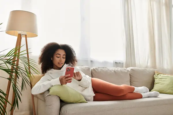 Une jeune femme aux cheveux bouclés profite de son temps à la maison, se prélassant sur le canapé tout en utilisant son téléphone. — Photo de stock