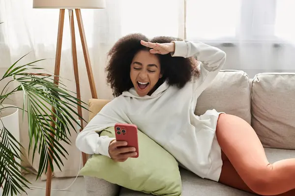 A young woman relaxes on her couch, playfully interacting with her phone while dressed comfortably. — Stock Photo