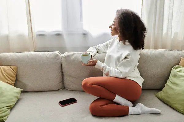 A young woman lounges comfortably, sipping a warm drink while enjoying the tranquility of her living room. — Stock Photo