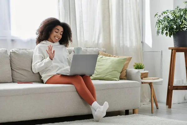 A cheerful woman in a cozy outfit joyfully connects with friends on her laptop from the couch. — Stock Photo