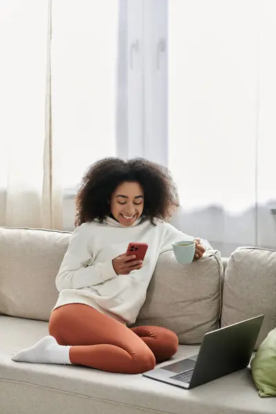 A young woman relaxes at home, sipping tea and using her phone while surrounded by comfort. — Stock Photo