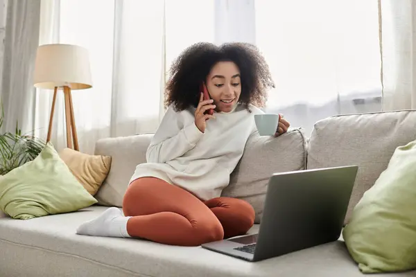 A joyful young woman relaxes at home, sipping tea while chatting on her phone and using her laptop. — Stock Photo