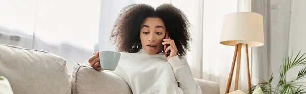 Une jeune femme se détend dans son salon, bavardant au téléphone tout en sirotant du thé. — Photo de stock
