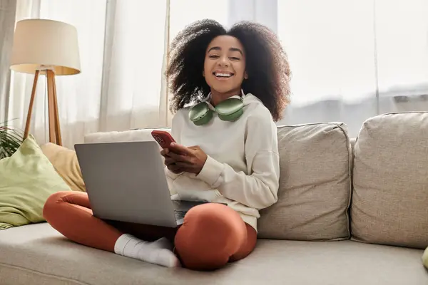 Una joven mujer se relaja en casa, sonriendo mientras usa su teléfono y su computadora portátil en un traje acogedor. - foto de stock