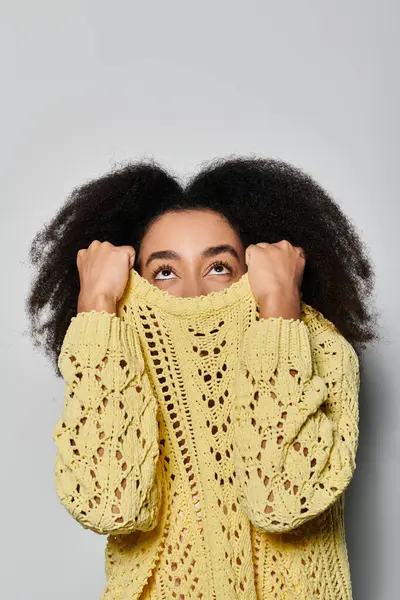 A young woman with curly hair playfully conceals her face in her yellow sweater, exuding charm. — Stock Photo