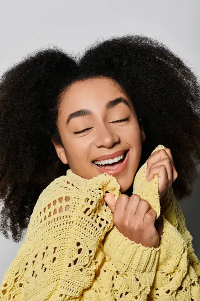 A radiant young woman smiles effortlessly while hugging her cozy yellow sweater in a serene studio. — Stock Photo