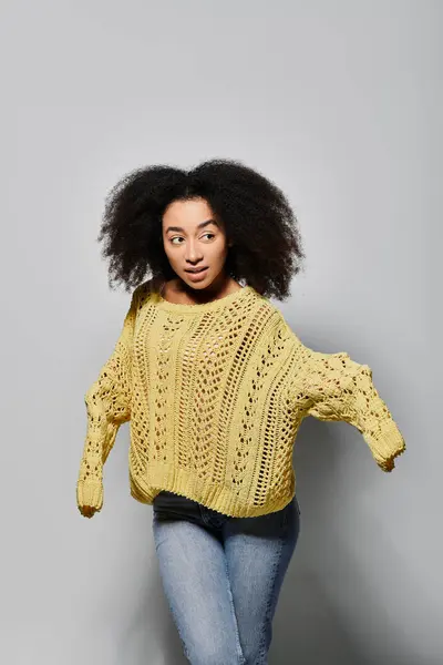 Una mujer joven con estilo en un suéter amarillo posa con confianza, destacando su cabello rizado sobre un telón de fondo neutro. - foto de stock