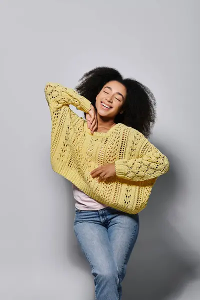 A cheerful young woman with curly hair dressed in a bright yellow sweater radiates joy against a grey backdrop. — Stock Photo