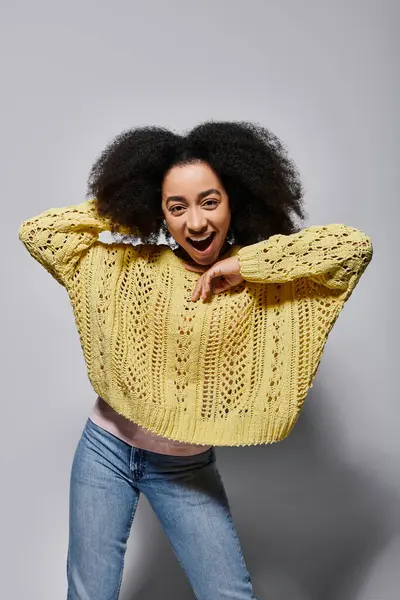 Eine junge Frau mit lockigem Haar posiert verspielt in einem gemütlichen gelben Pullover, der Freude und Zuversicht ausstrahlt.. — Stockfoto