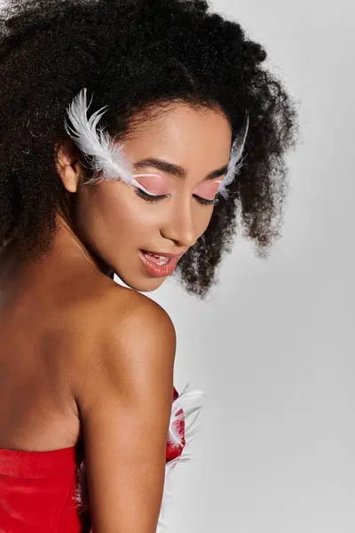 A young woman with curly hair elegantly showcases her vibrant red outfit, adorned with feathers, on a subtle grey background. — Stock Photo