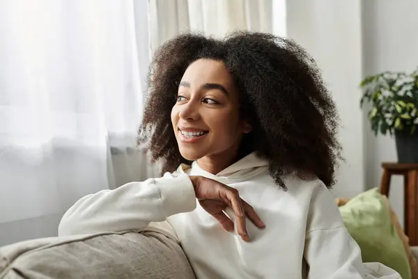 A young woman in a cozy hoodie sits comfortably, enjoying her time in a sunny living room. — Stock Photo