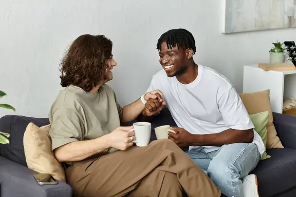 Two men share laughter and love while sipping coffee on a cozy couch in their home. — Stock Photo