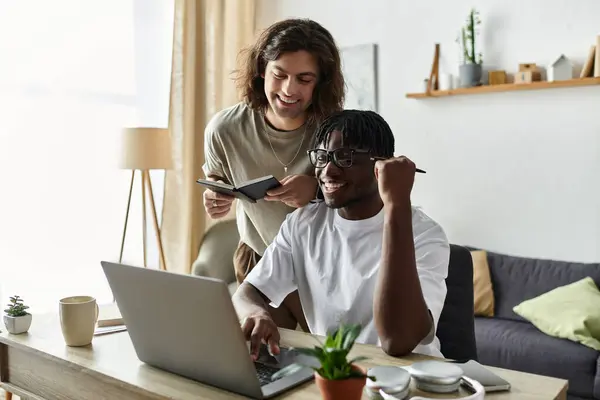 Dos hombres comparten un momento amoroso en casa, uno felizmente ayudando al otro con una tarea de computadora portátil. - foto de stock