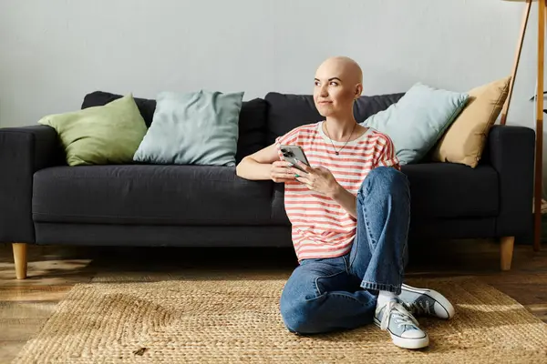 Una mujer calva se relaja en el suelo, disfrutando de su teléfono en medio de un espacio cálido y acogedor. - foto de stock