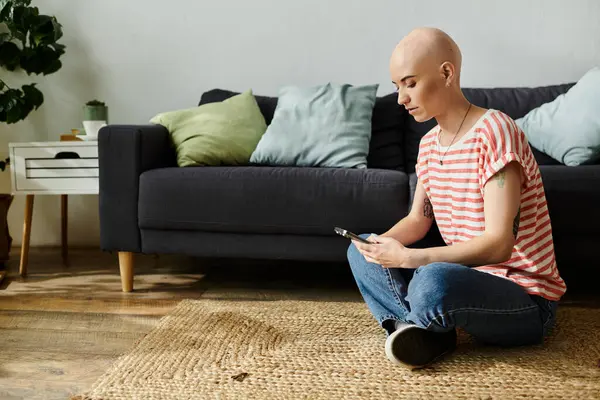 A young bald woman sits cross legged on a rug, focused on her phone in a cozy room. — Stock Photo